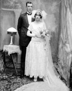 A black and white 1920s wedding portrait photograph, with the tall man standing behind the short woman, with a gap between them.
